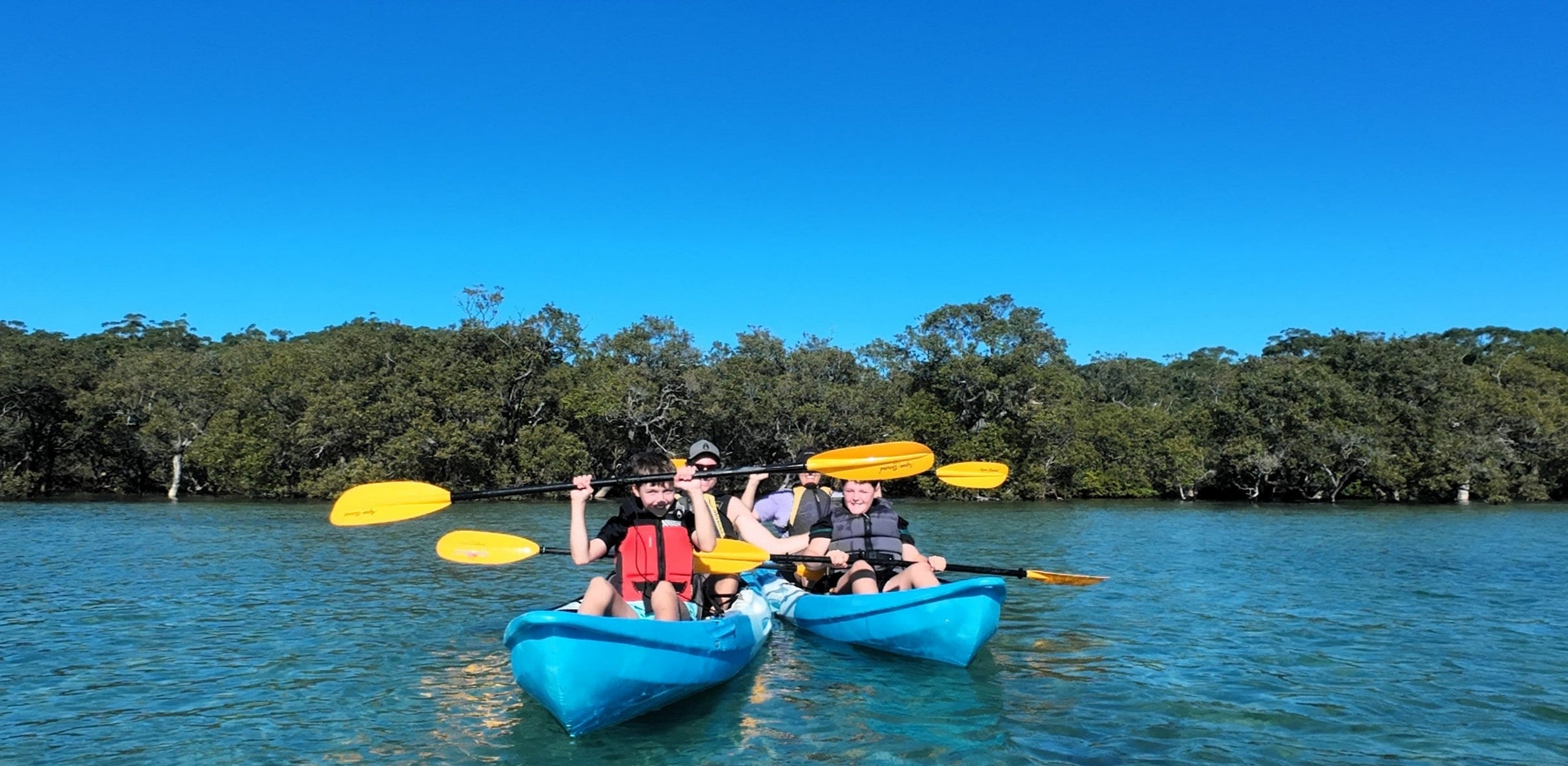 Two people in blue kayaks paddling on a clear body of water with trees in the background.